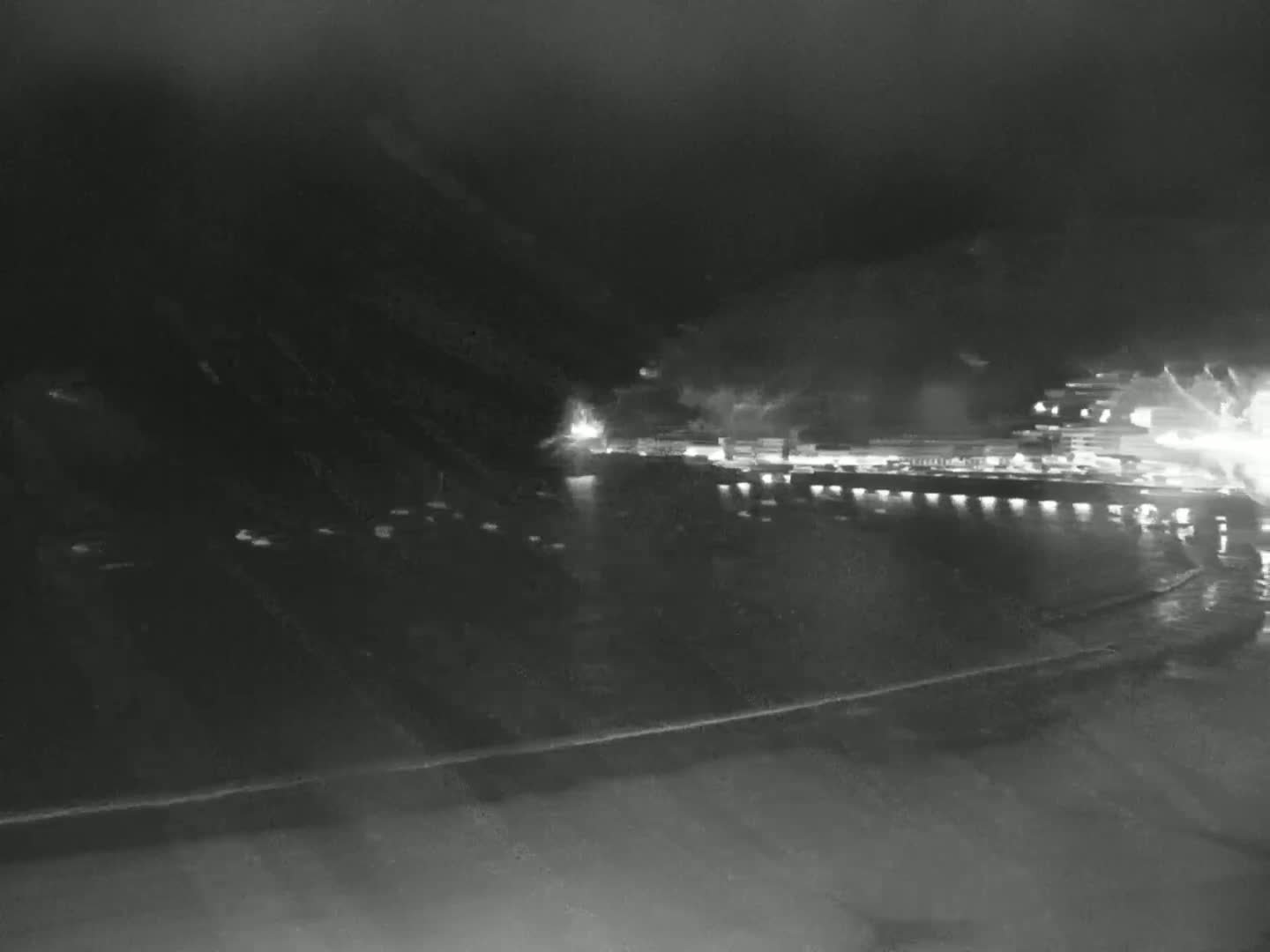 A nighttime view of a dimly lit waterfront, with a dark hillside rising behind it.