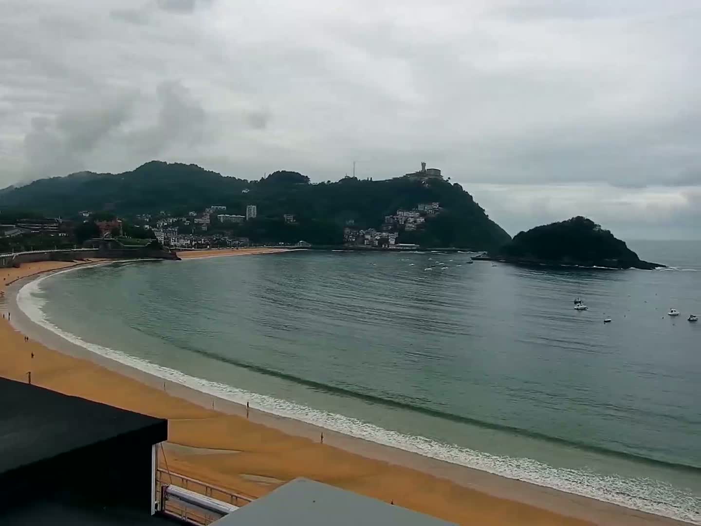 A curving sandy beach and calm, grey-green ocean are nestled beneath a cloudy sky, with a town and a hill rising in the background.