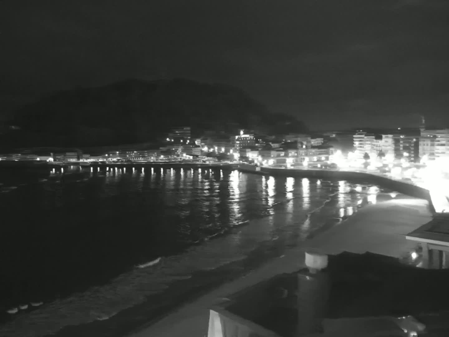 A nighttime view of a town's illuminated waterfront, with calm waves lapping a sandy beach under a dark sky.