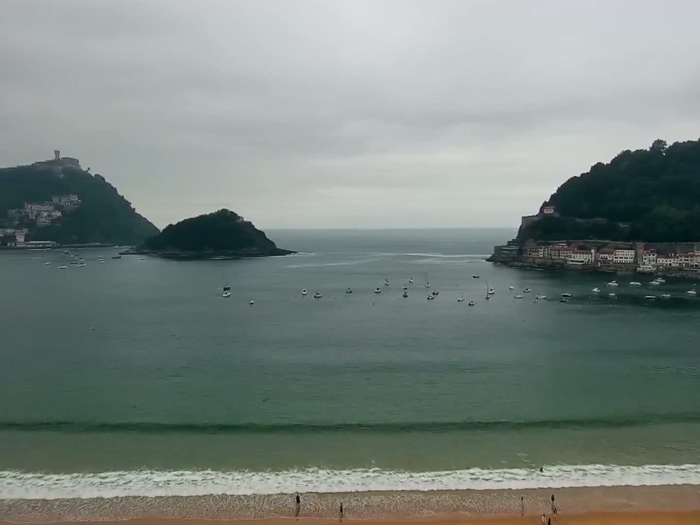 A sandy beach with gentle waves meets a calm bay dotted with boats, nestled between two verdant hills under an overcast sky.
