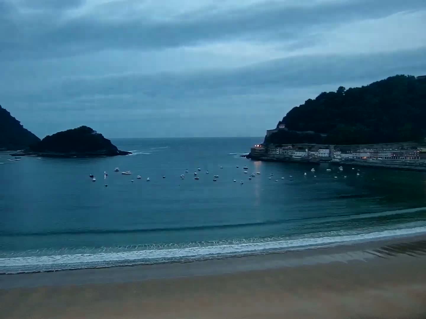 A calm, dark-blue bay with several small boats is nestled between two hills, one of which has buildings along its shore, under a cloudy sky.