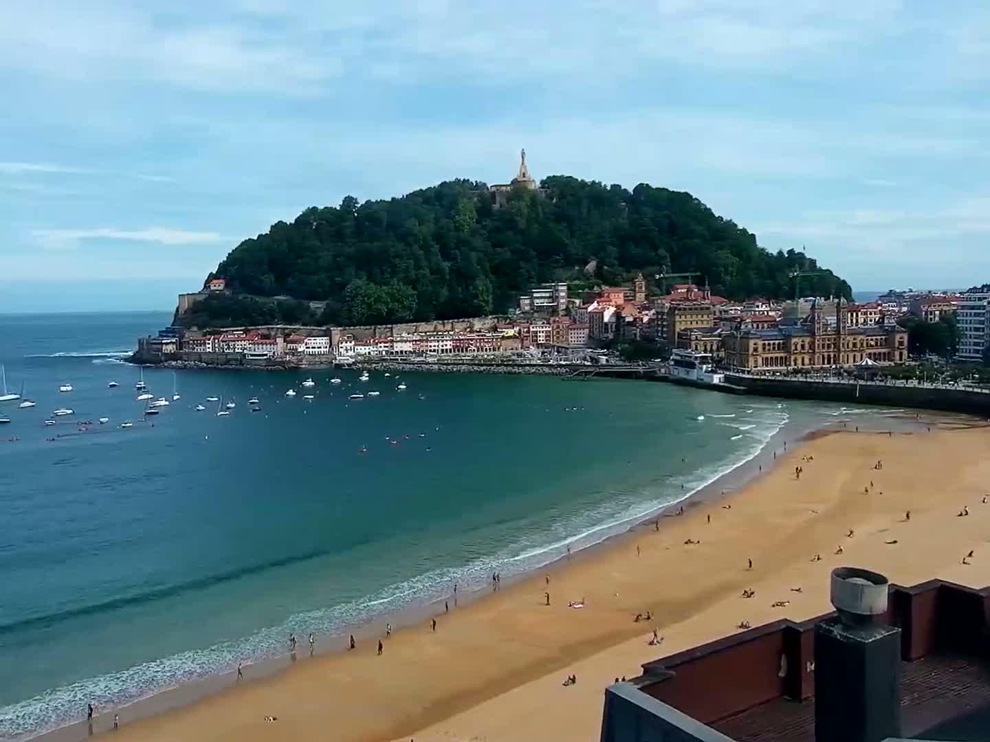 A sandy beach with people sunbathing curves around a calm bay, in front of a town nestled at the base of a green hill topped with a monument, under a mostly clear sky.