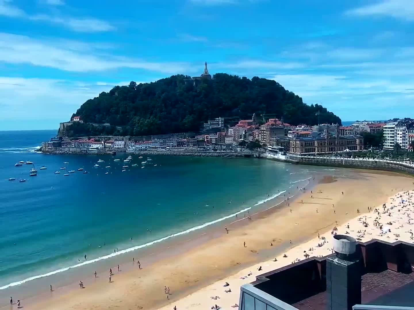A sandy beach with scattered sunbathers curves around a bay, facing a town nestled at the base of a verdant hill under a clear, bright blue sky.