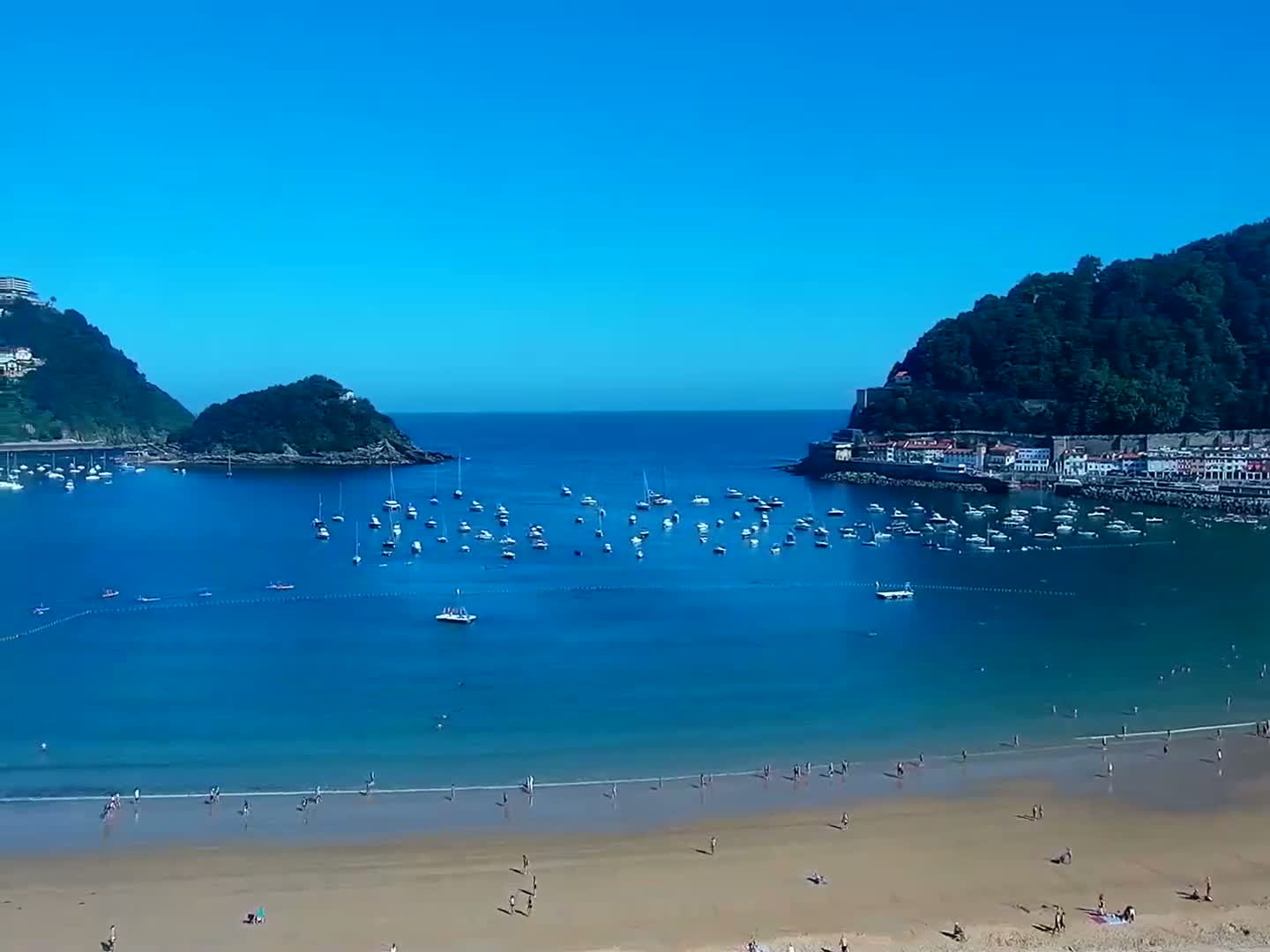 A sandy beach with many people on it is nestled in a calm, blue bay surrounded by green hills and numerous boats under a clear blue sky.