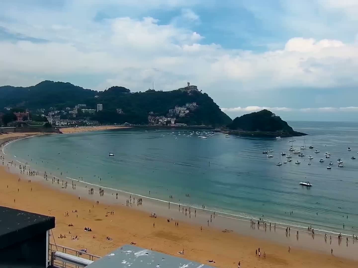A sandy beach with many people on it is nestled in a calm, blue bay surrounded by green hills and numerous boats under a clear blue sky.