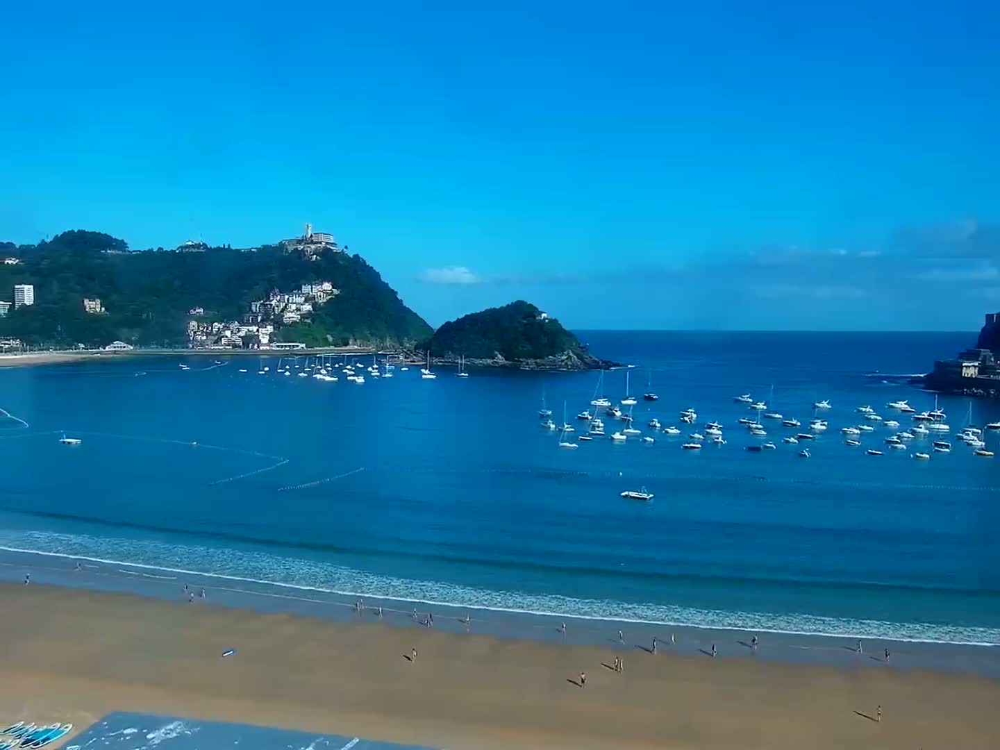 A sandy beach with a calm ocean and numerous boats in a bay, backed by green hills and a clear blue sky.