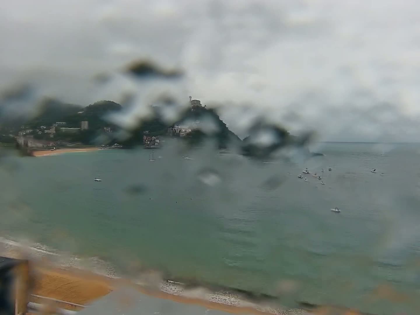 A rain-streaked view shows a bay with a sandy beach, several small boats, and a hilltop town nestled against a mountain range under an overcast sky.