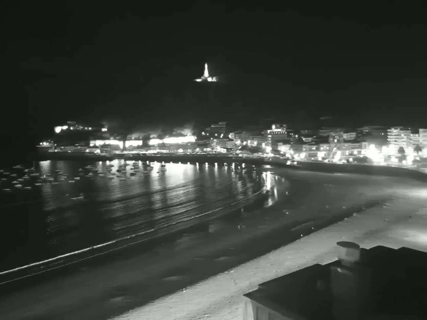 A nighttime view of a calm bay with a sandy beach, a town with lights, and a hilltop structure in the distance.