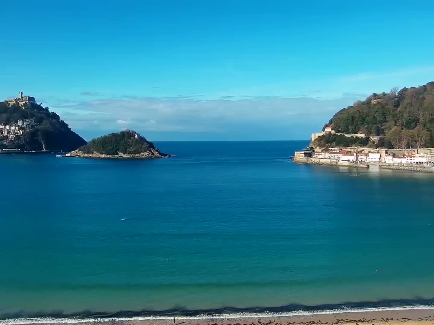 A nighttime view of a calm bay with a sandy beach, a town with lights, and a hilltop structure in the distance.