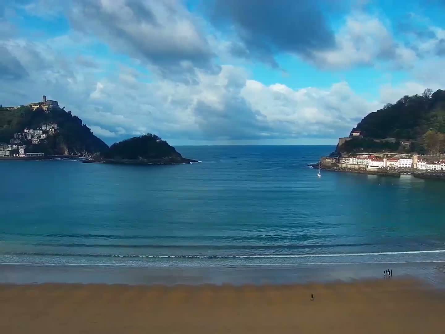 A scenic bay with a wide sandy beach in the foreground is bordered by urbanized hills on both sides, an island in the middle, and a distinctive fortress atop the left hill, all under a partly cloudy sky.