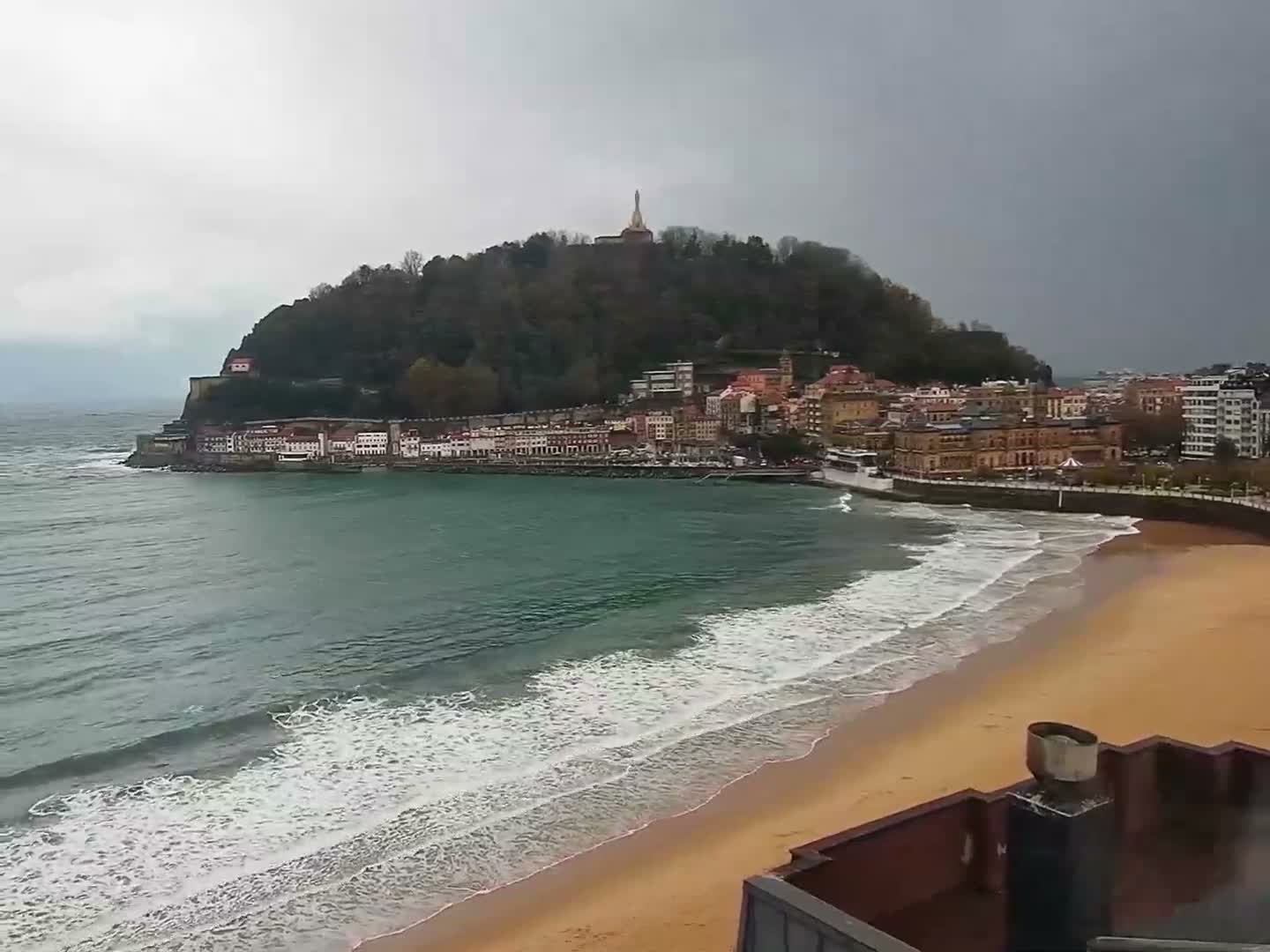 An elevated view captures a vibrant coastal city with colorful buildings lining a sandy beach and a bay featuring breaking waves, all set against a verdant mountain topped with a prominent statue, under a heavily overcast and gloomy sky.