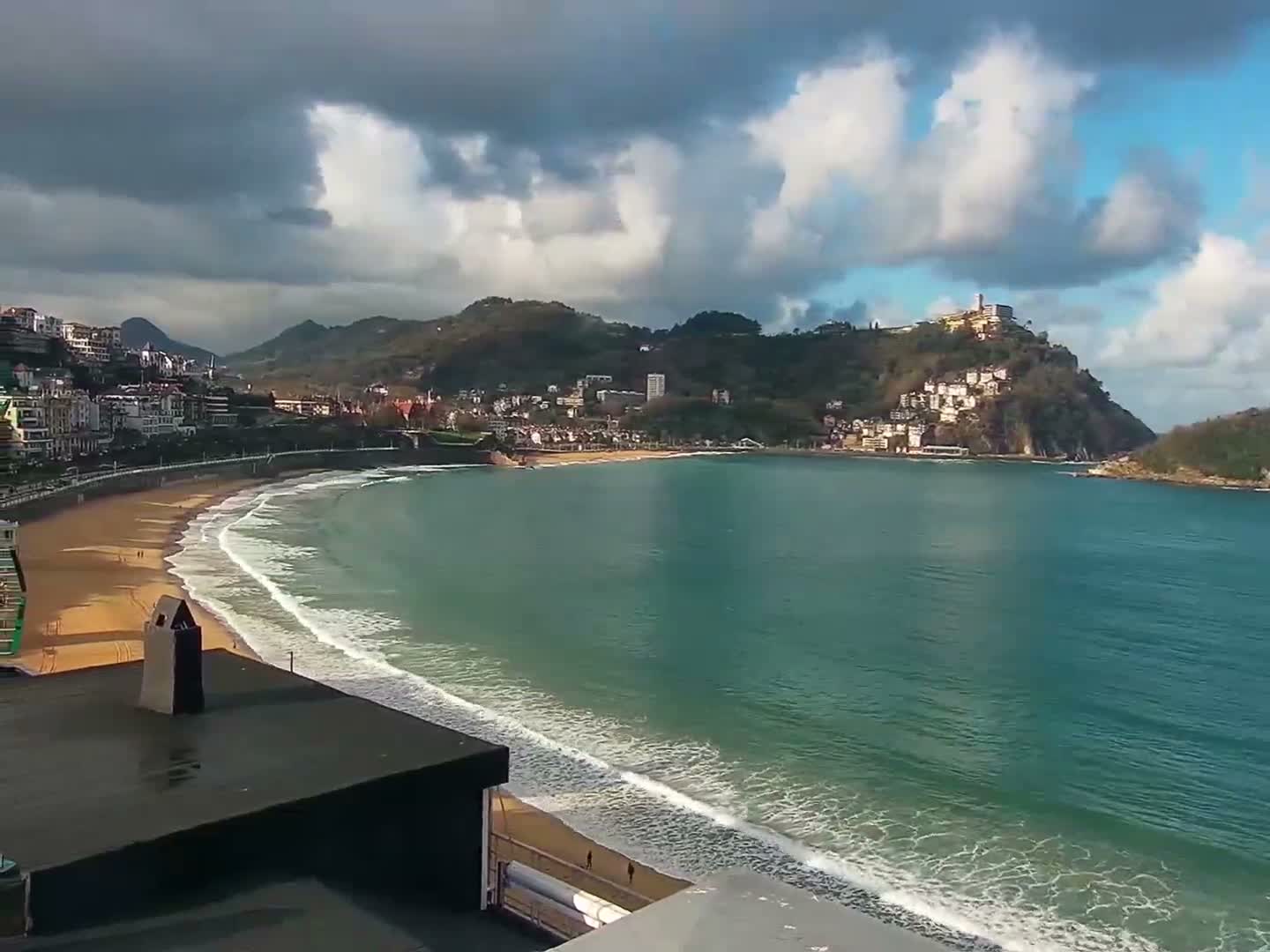 A panoramic view reveals a wide bay with a curving sandy beach and gentle waves, flanked by a city nestled against green mountains and a prominent hill topped with a fortress, all beneath a partly cloudy sky with dramatic dark clouds and bright blue patches.
