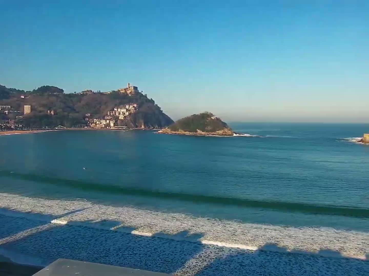 A panoramic view reveals a wide bay with a curving sandy beach and gentle waves, flanked by a city nestled against green mountains and a prominent hill topped with a fortress, all beneath a partly cloudy sky with dramatic dark clouds and bright blue patches.
