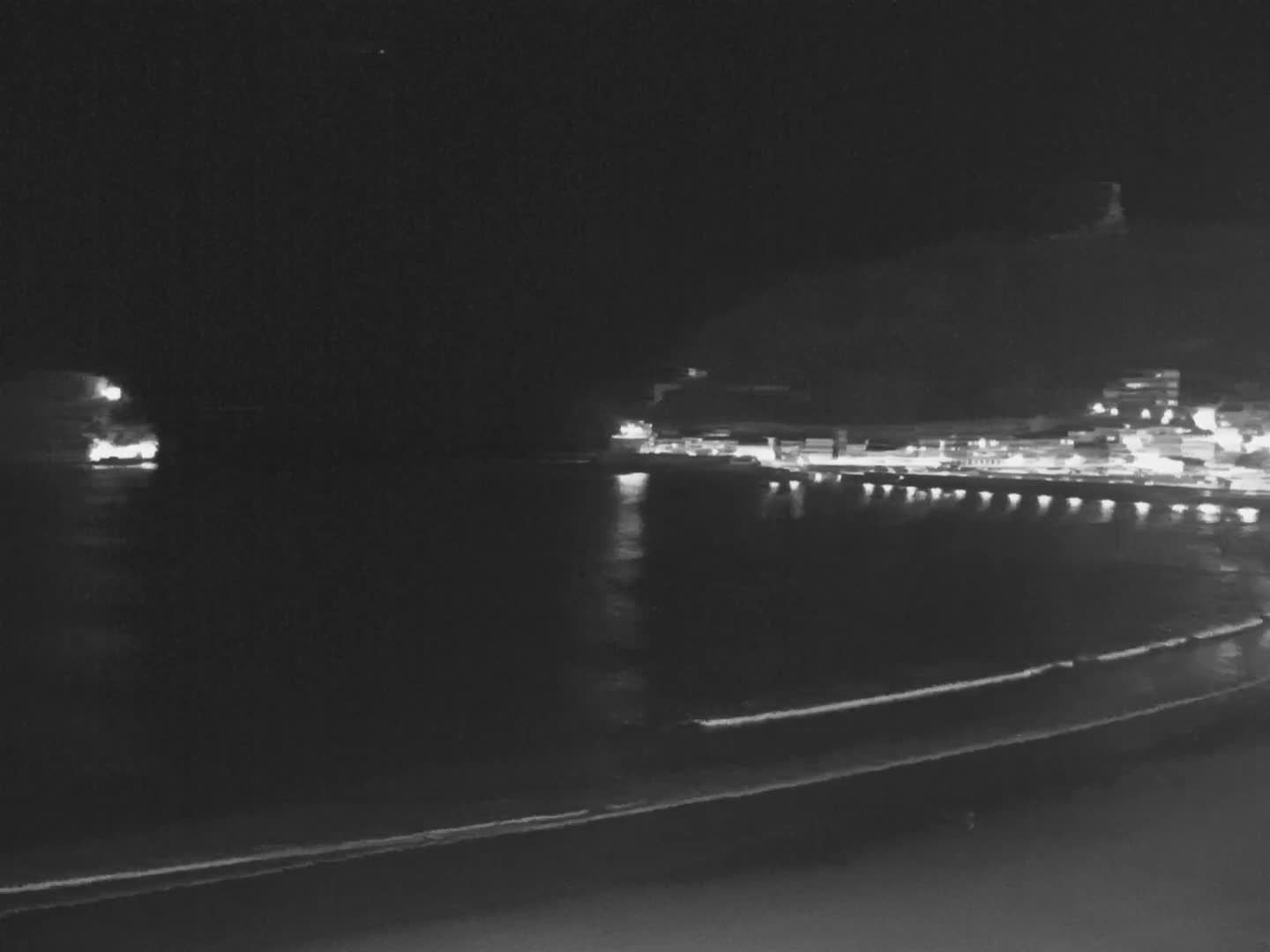 A panoramic view reveals a wide bay with a curving sandy beach and gentle waves, flanked by a city nestled against green mountains and a prominent hill topped with a fortress, all beneath a partly cloudy sky with dramatic dark clouds and bright blue patches.