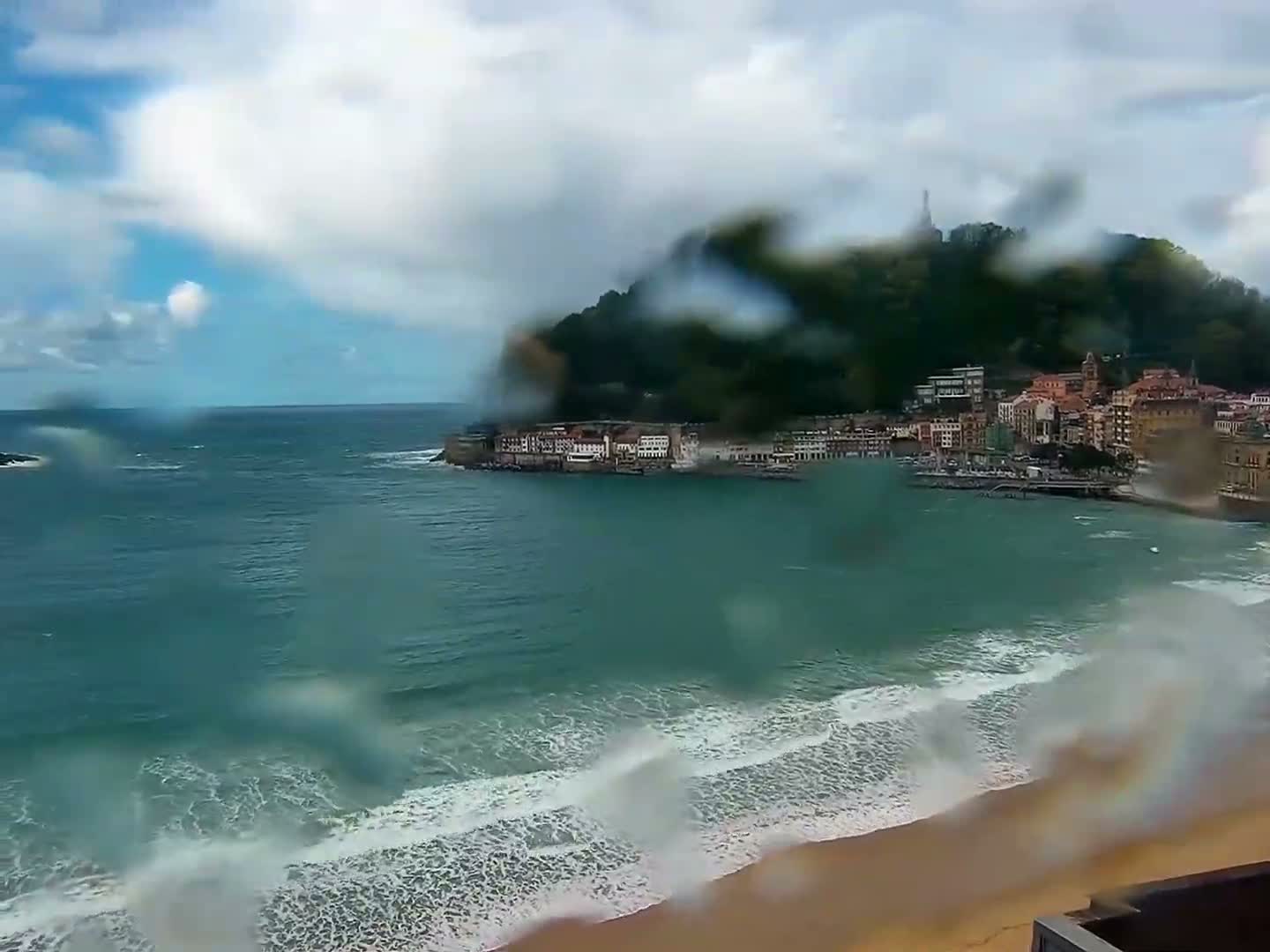A panoramic view reveals a wide bay with a curving sandy beach and gentle waves, flanked by a city nestled against green mountains and a prominent hill topped with a fortress, all beneath a partly cloudy sky with dramatic dark clouds and bright blue patches.