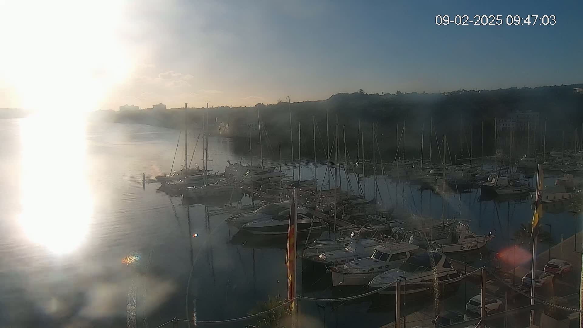 A bright sunrise illuminates a calm harbor filled with numerous docked sailboats and motorboats, partially obscured by a hazy atmosphere.