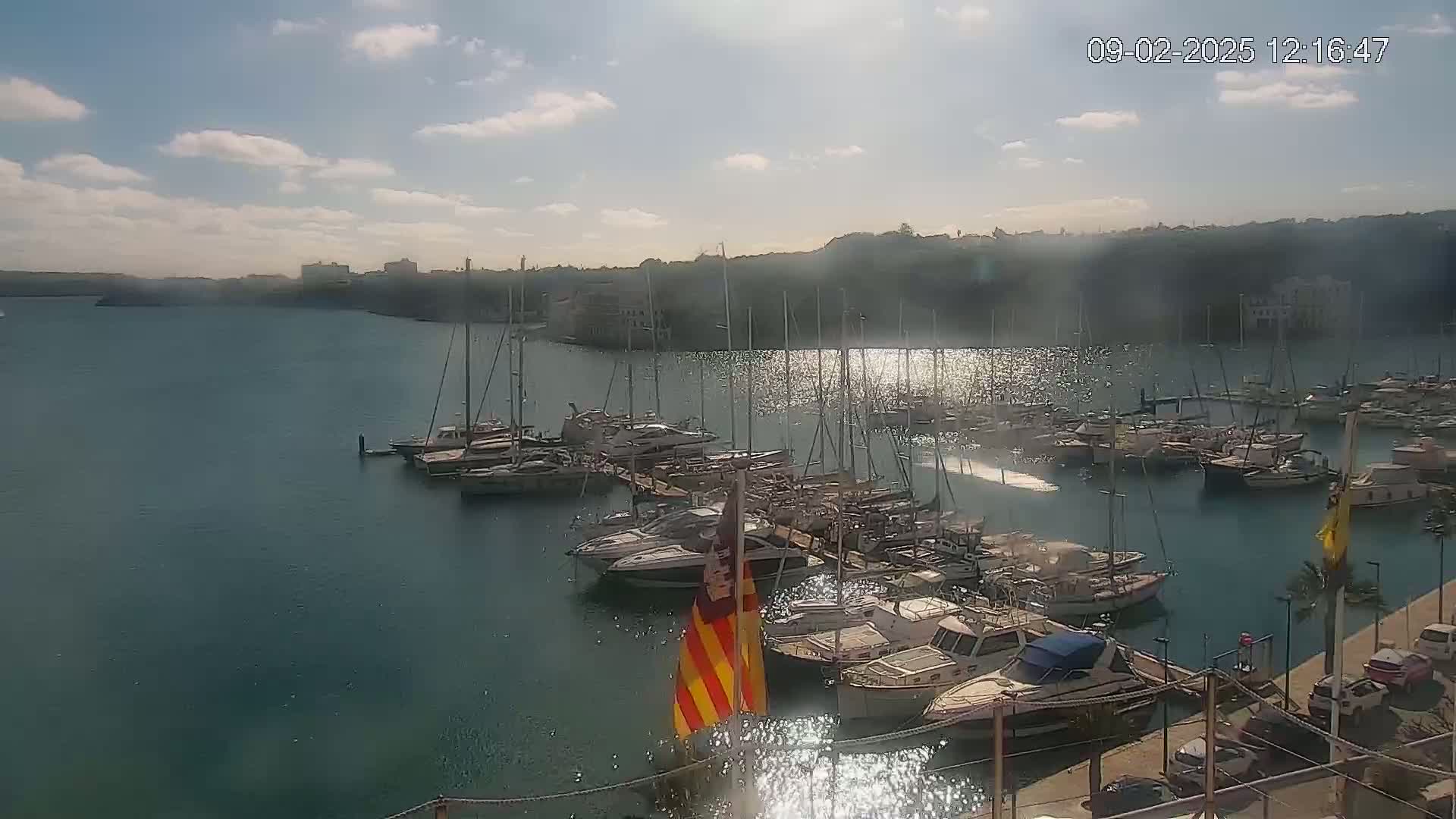 A marina filled with numerous yachts and sailboats is situated on calm, sparkling water under a mostly sunny sky.