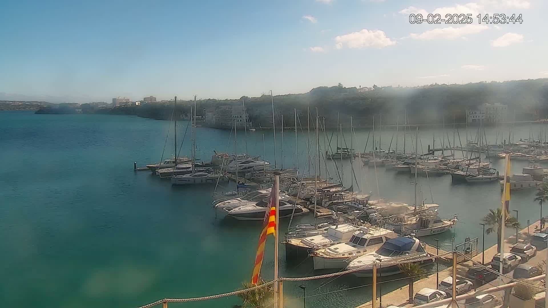 A marina filled with numerous yachts and sailboats is situated on calm, teal water under a mostly sunny sky with a few scattered clouds.
