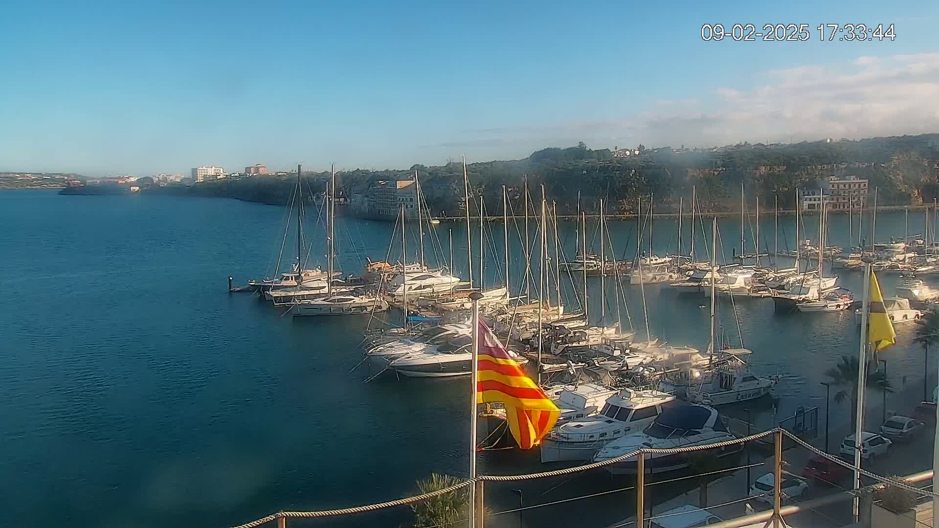A calm, sunny day reveals a marina filled with numerous sailboats and motorboats, nestled in a bay next to a town on a hillside.