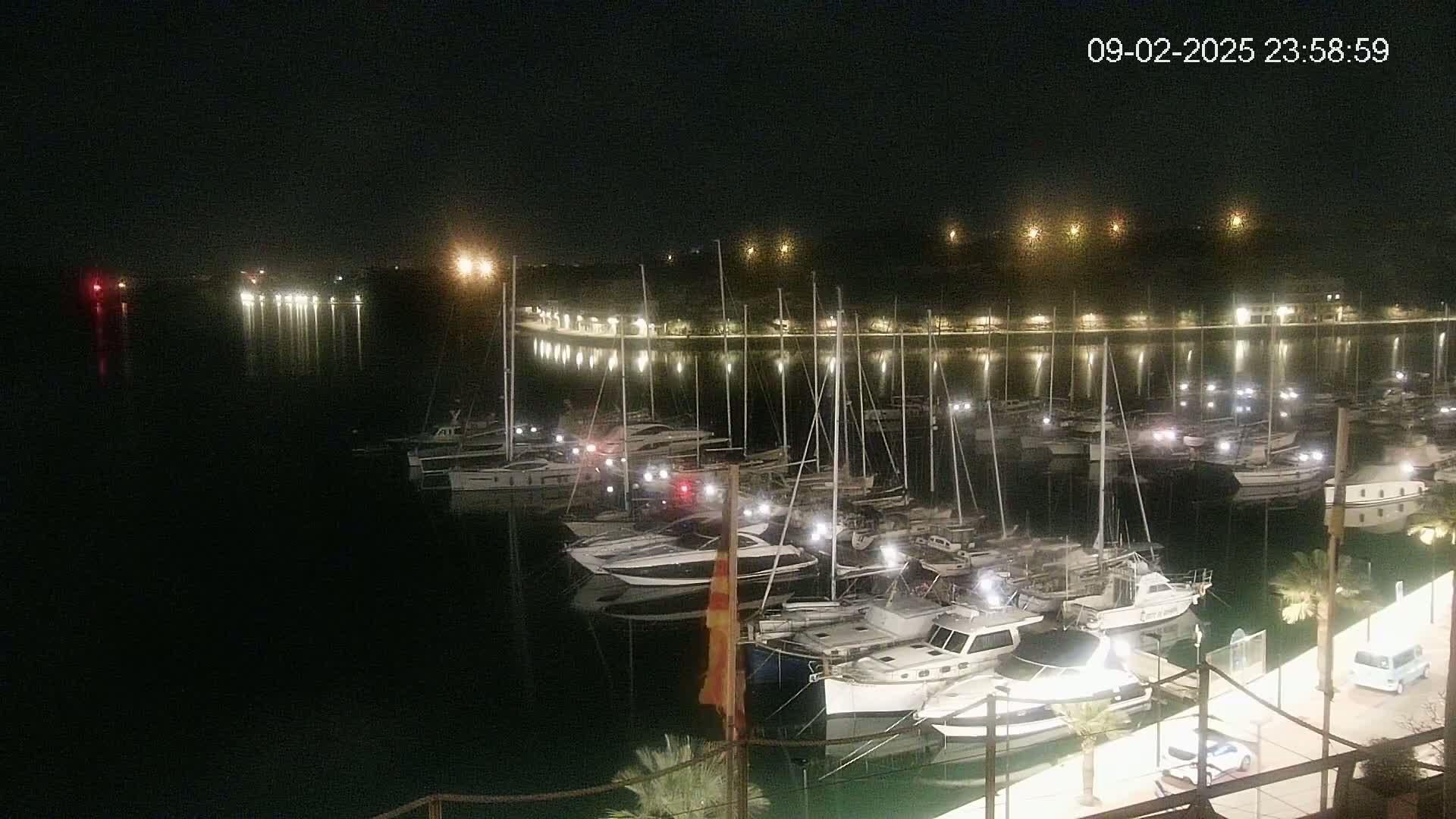 At night, many boats are docked at a marina, illuminated by lights along the shoreline reflecting on calm water.