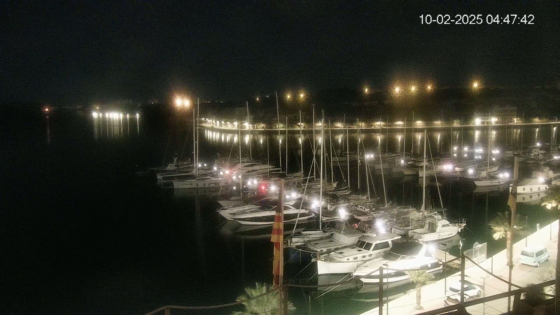 Many boats are docked at a marina at night, illuminated by lights along the shoreline.