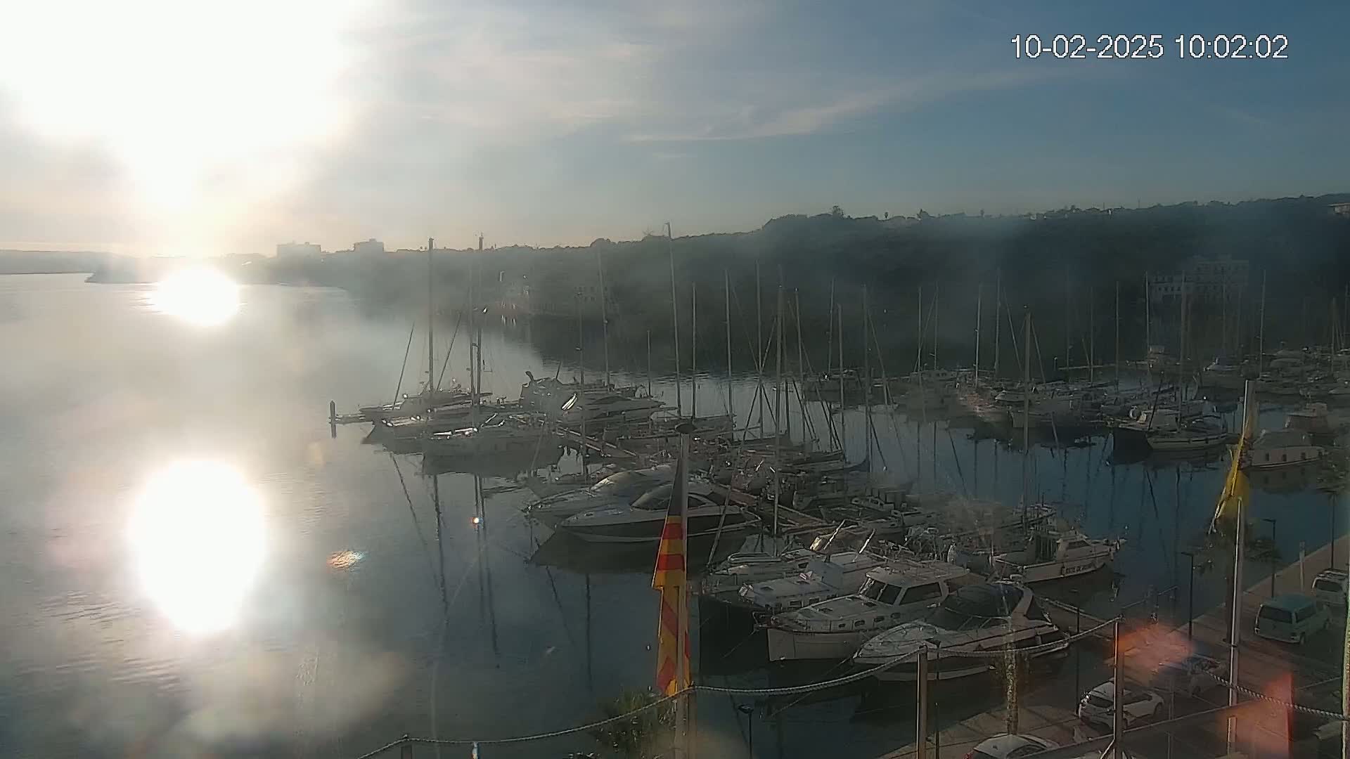 A marina filled with numerous sailboats and motorboats is seen at sunrise on a partly sunny day with a light haze over the water.