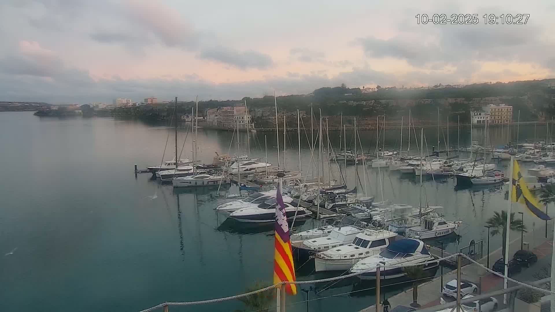 A marina filled with numerous sailboats and motorboats is situated on calm water under a cloudy, dusky sky.