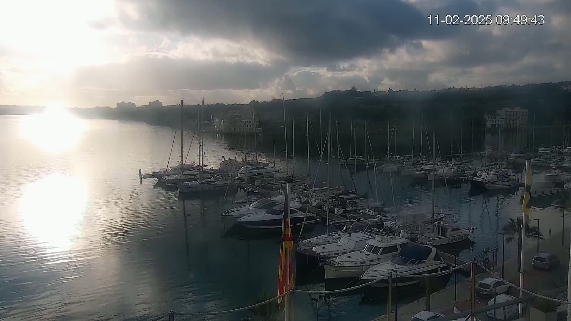 A calm, sunlit harbor filled with numerous yachts and sailboats under a partly cloudy sky.