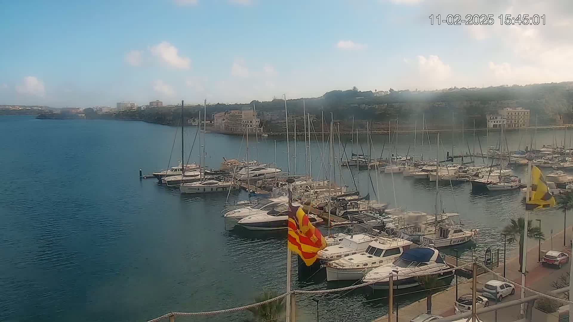 A marina filled with numerous yachts and sailboats is situated on calm, blue water under a mostly clear sky with a few scattered clouds.