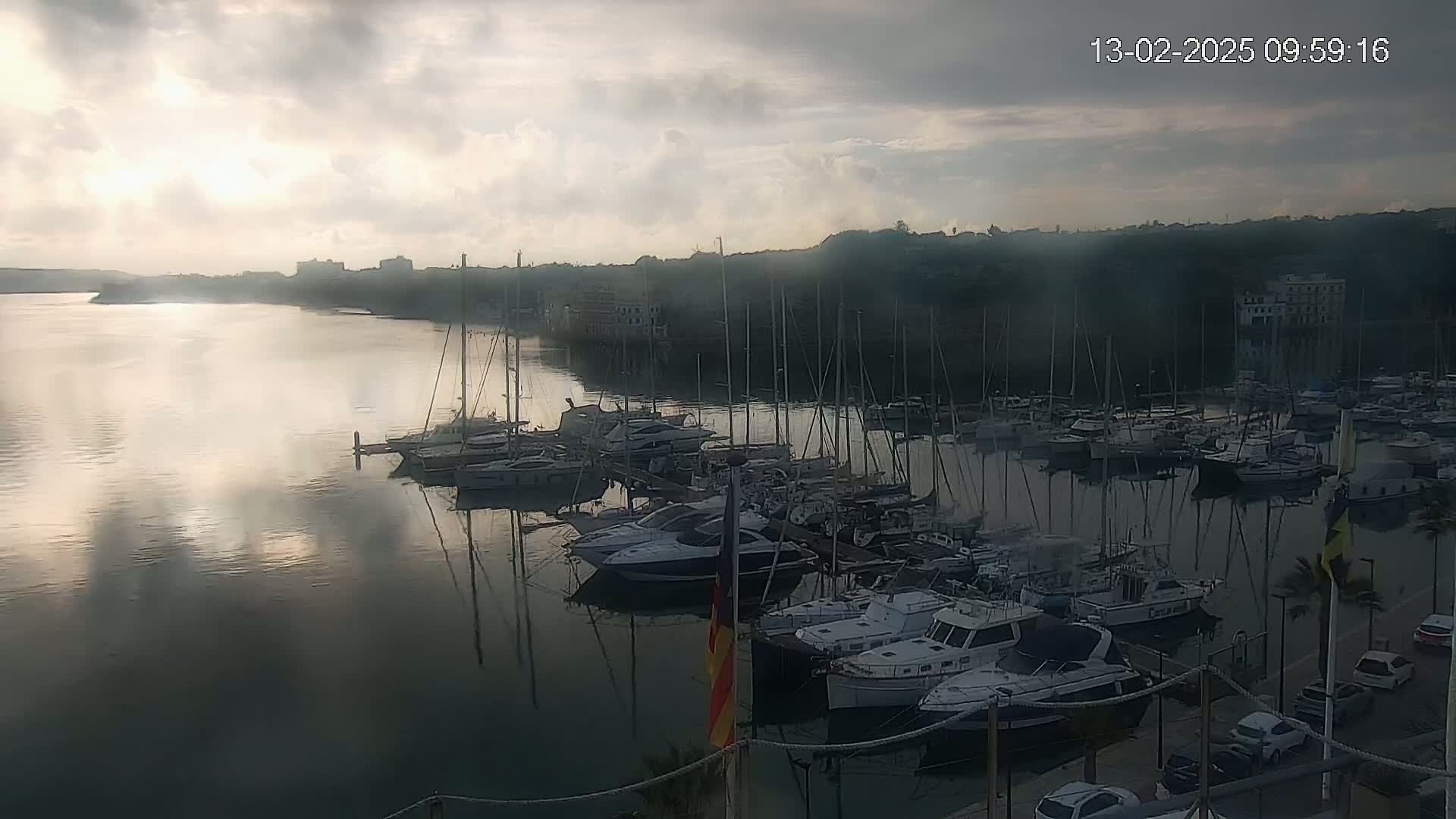 Many sailboats and motorboats are docked in a calm harbor under a cloudy but bright sky.