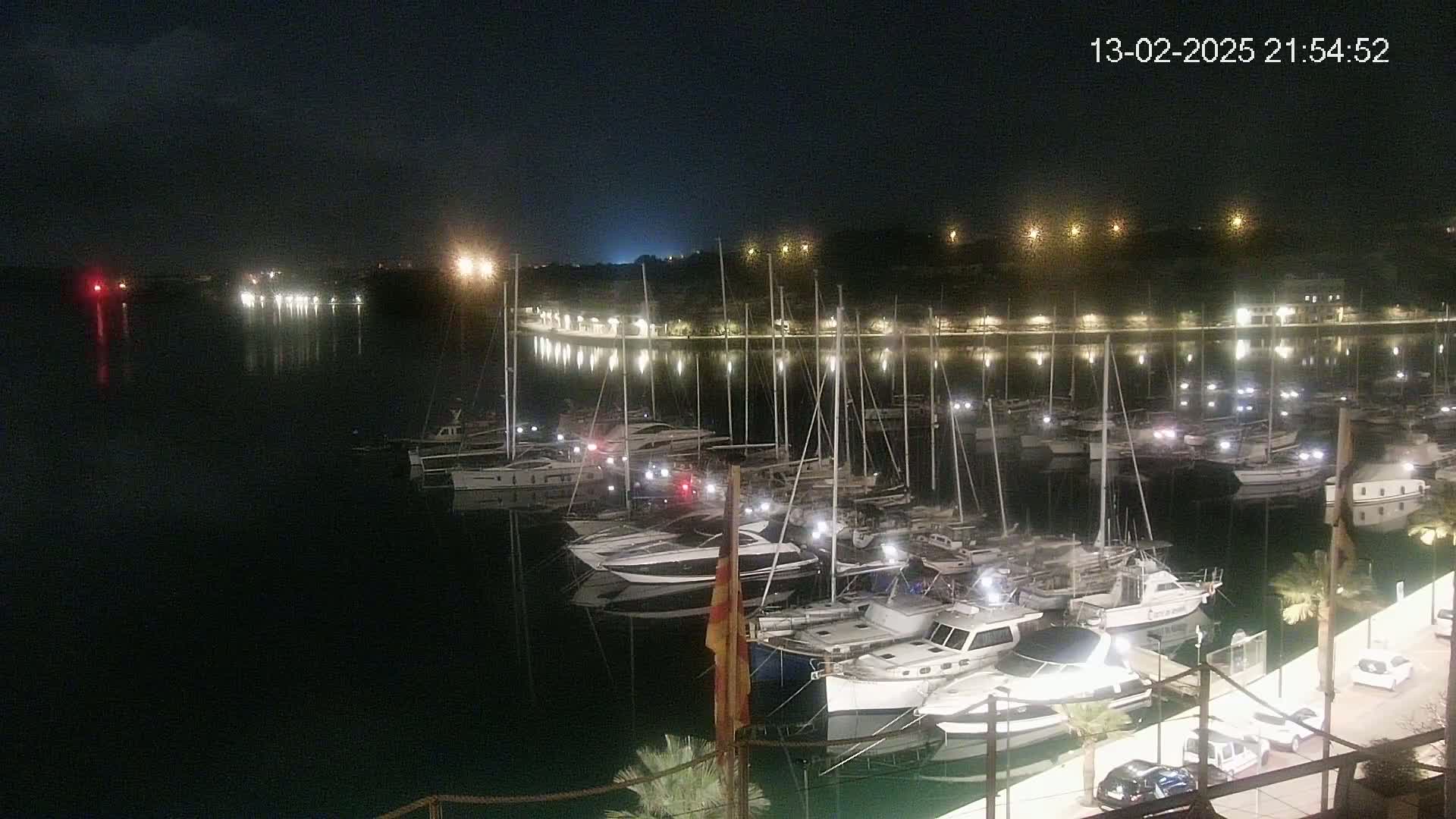 Many boats are docked in a marina at night, illuminated by lights along the shore and on the boats themselves.