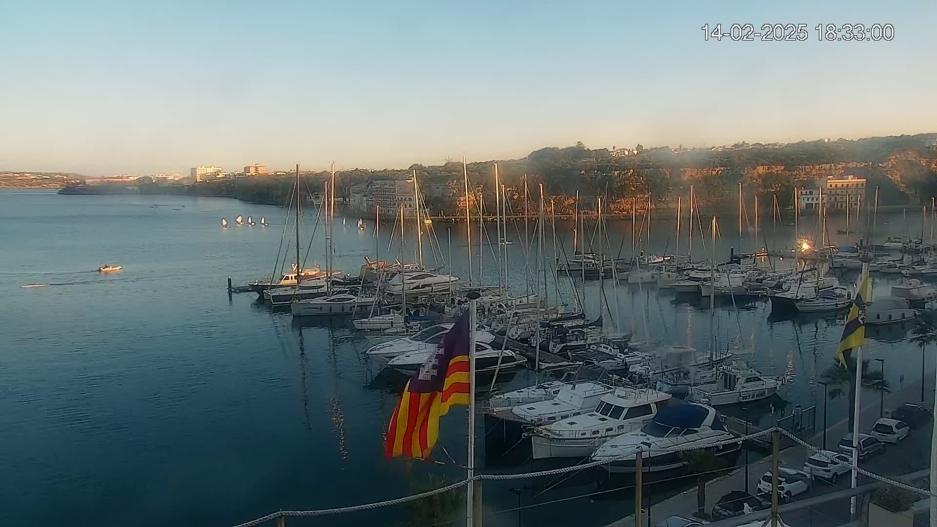 A calm, sunlit harbor filled with sailboats and motorboats, with a few small sailboats visible in the distance.