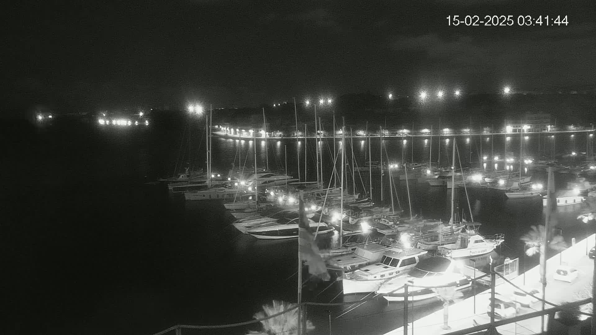 A nighttime, black-and-white view of a marina filled with numerous boats and yachts, illuminated by lights along the docks.