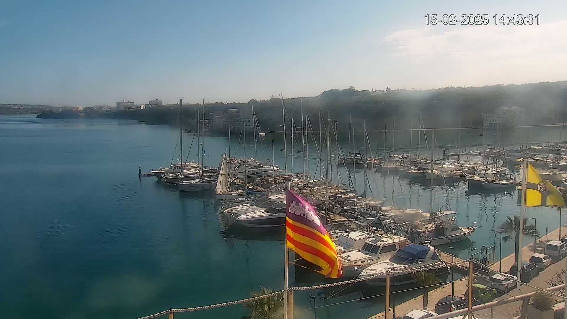 A marina filled with numerous sailboats and motorboats is situated on calm, clear blue water, under a mostly sunny sky with a light haze.