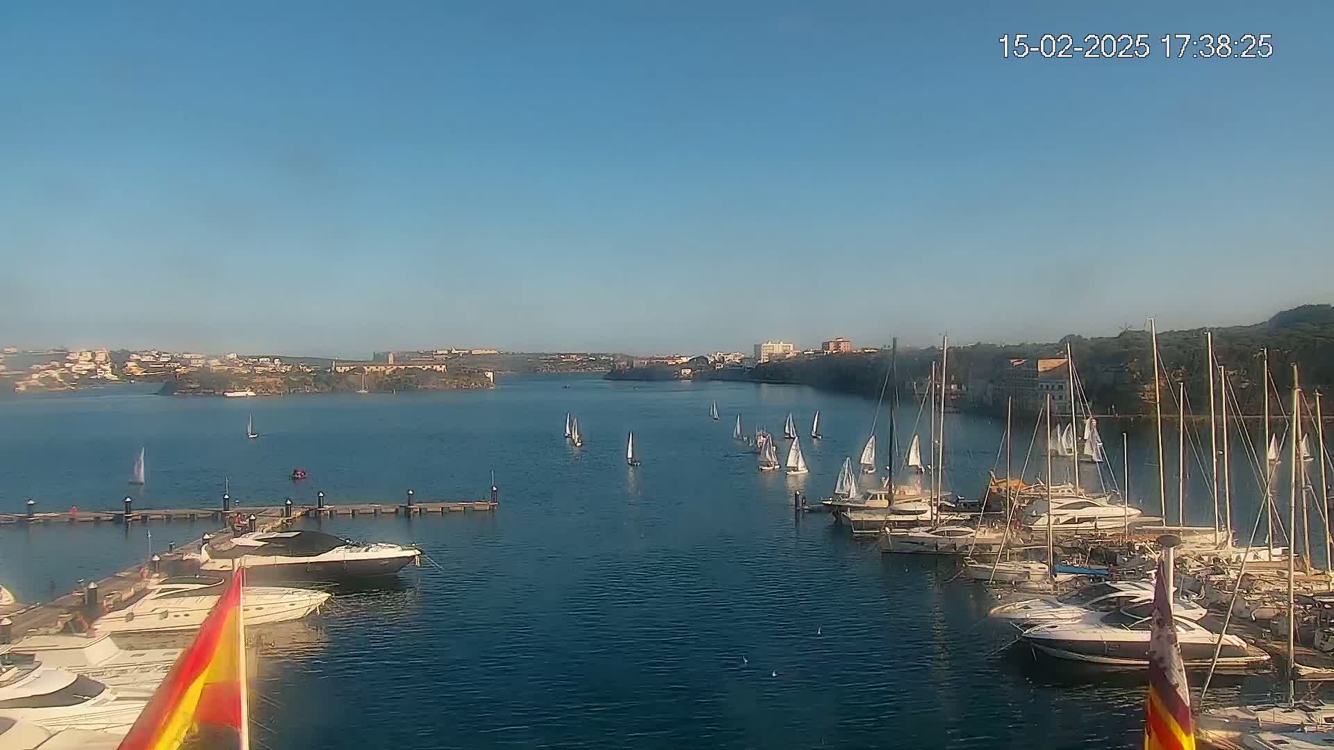 Under a clear blue sky, numerous sailboats are scattered across a calm bay, with many more docked in a marina filled with motor yachts.