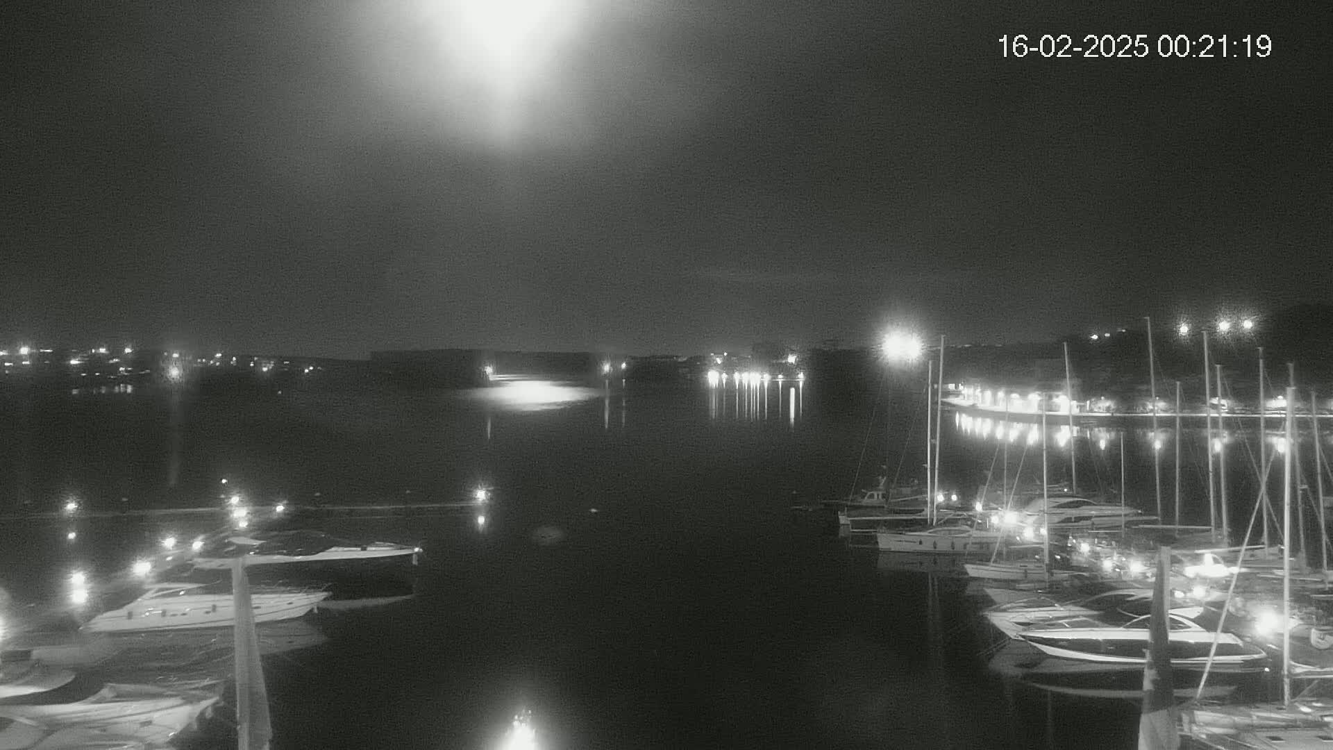 A nighttime, low-light view of a harbor filled with boats, illuminated by lights along the shore under a bright moon.
