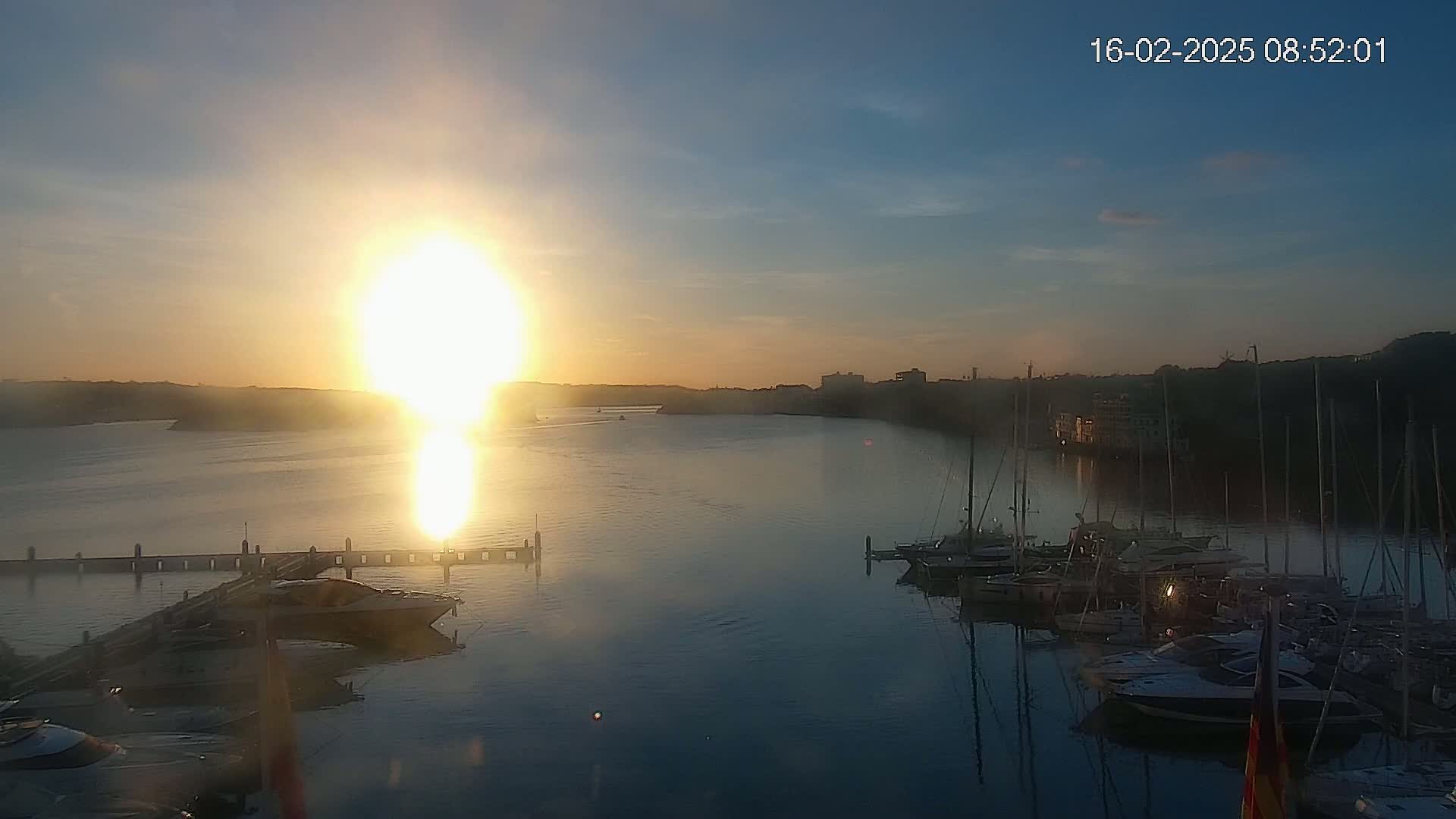 A bright sunrise over a calm bay filled with sailboats docked at a marina.