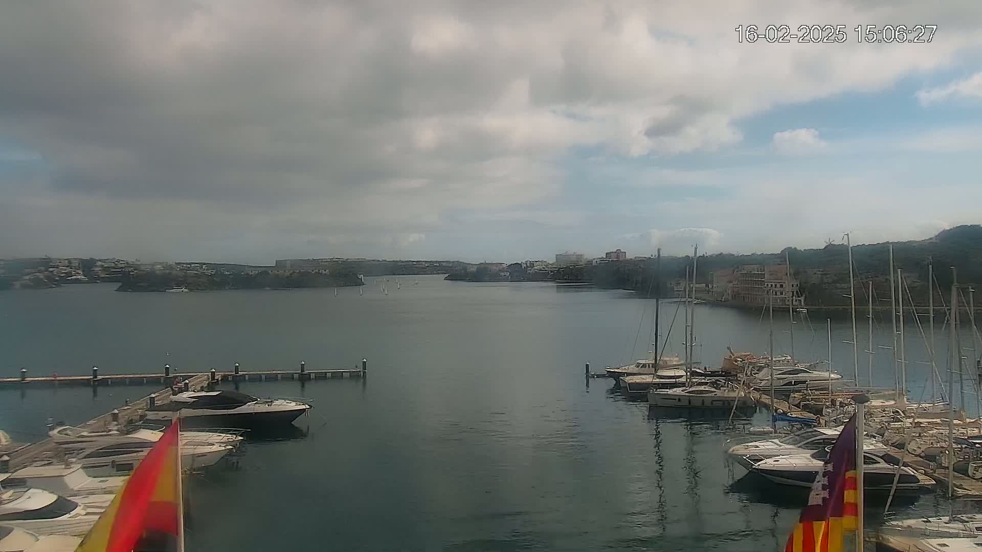 A calm bay with overcast skies features numerous yachts and sailboats docked at marinas, with a town visible in the distance.