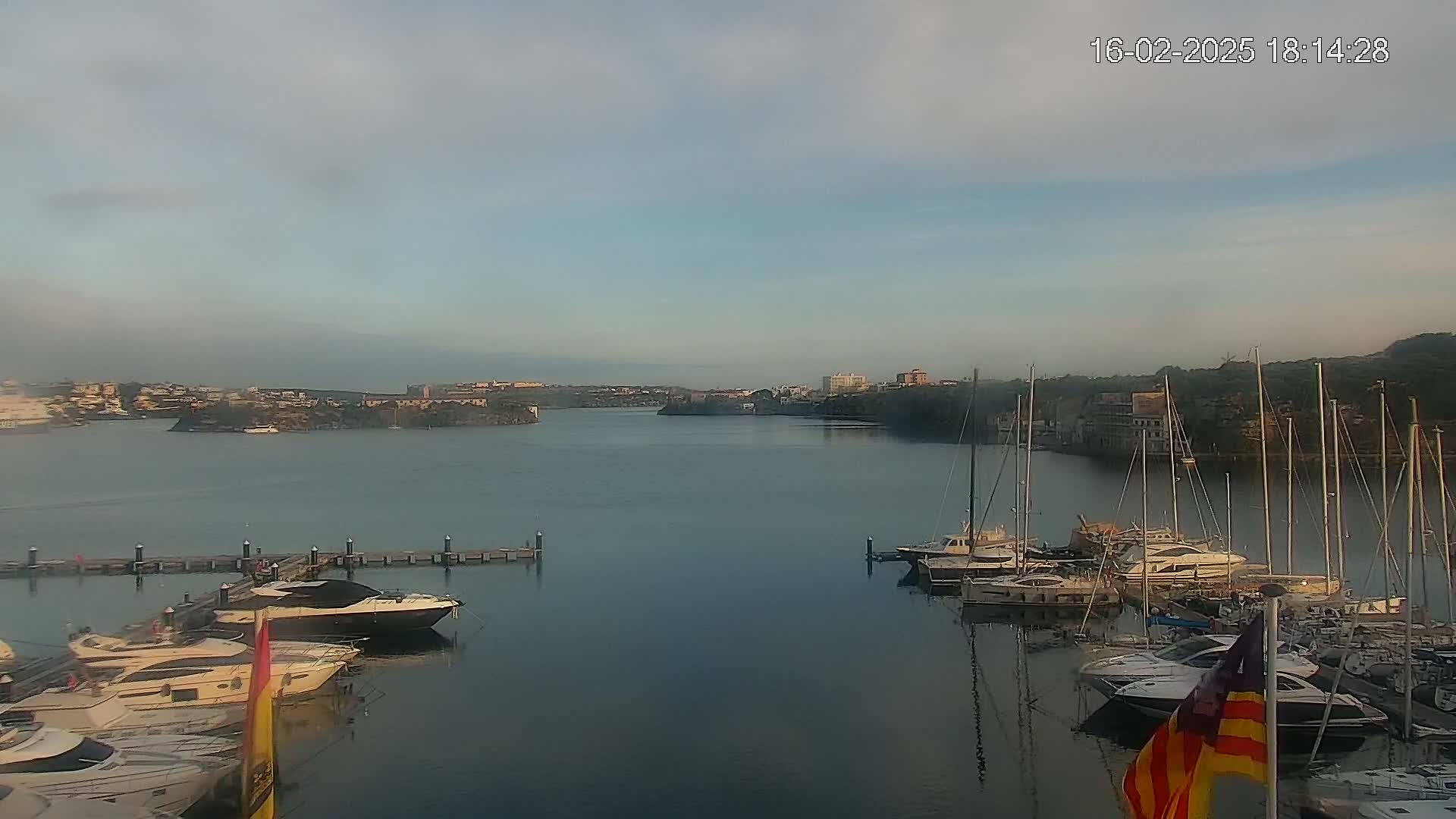 A calm bay with numerous yachts and sailboats docked at a marina, under a hazy, overcast sky.