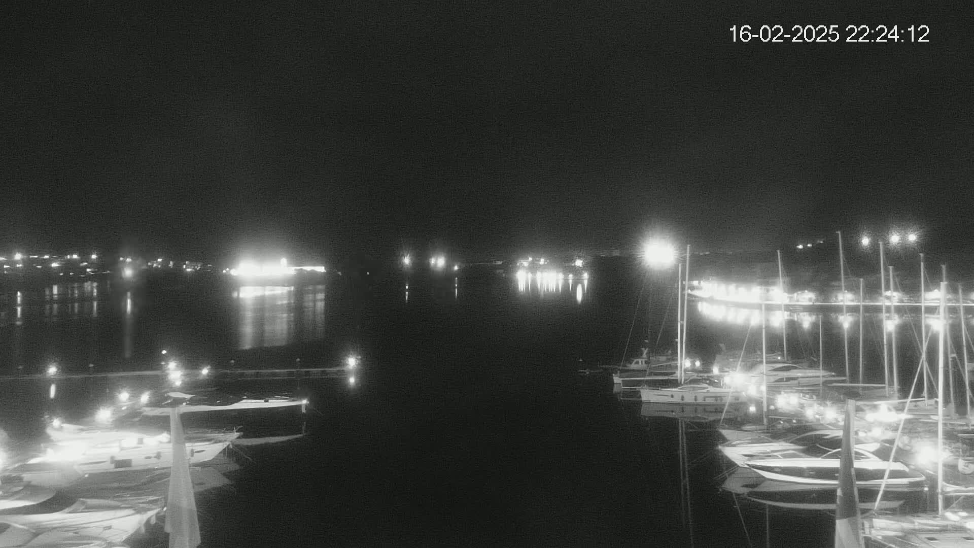A nighttime view of a marina with many boats docked, illuminated by lights, across a calm body of water under a dark sky.