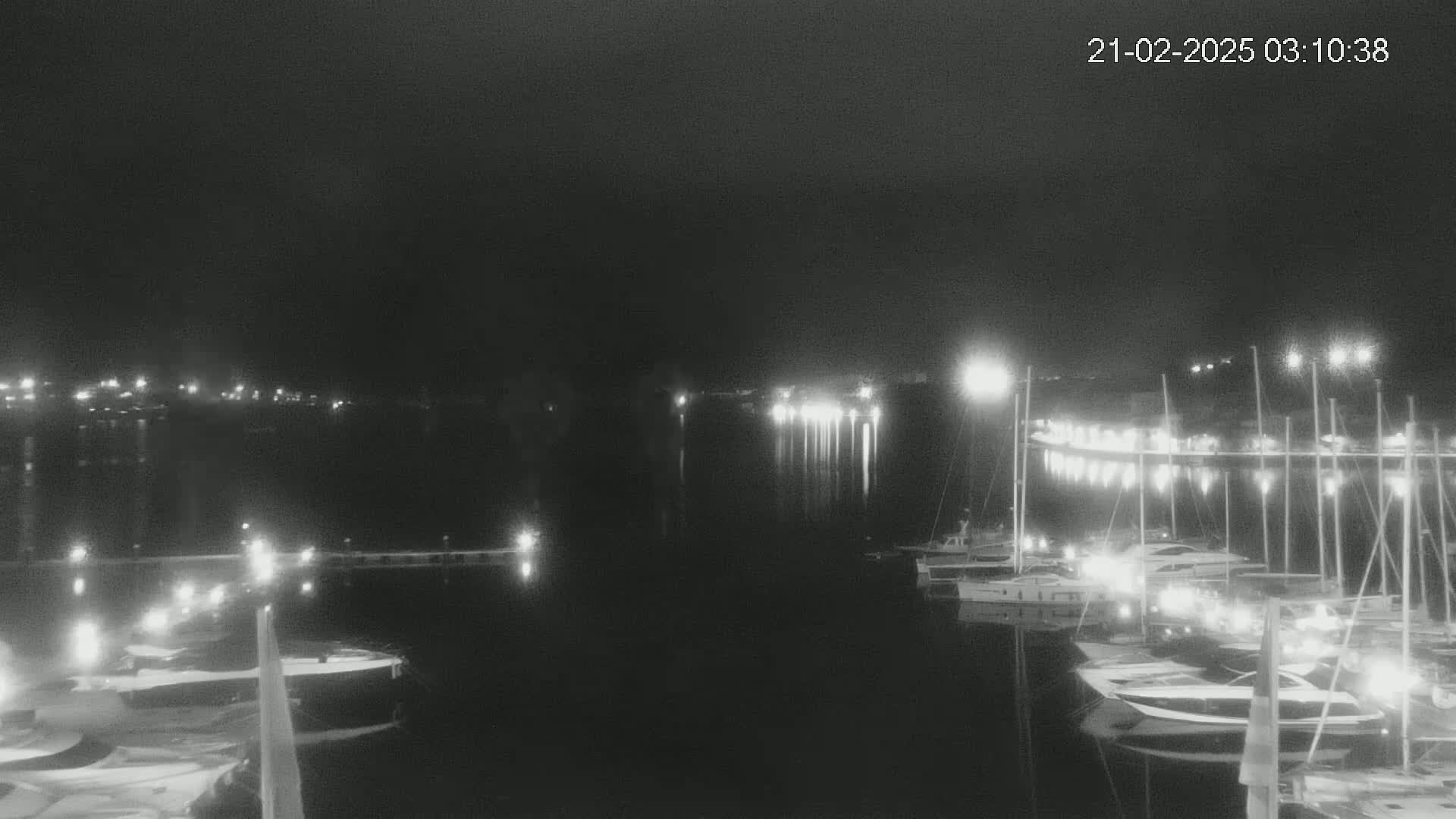 A nighttime view of a harbor filled with boats illuminated by lights under a dark, cloudy sky.