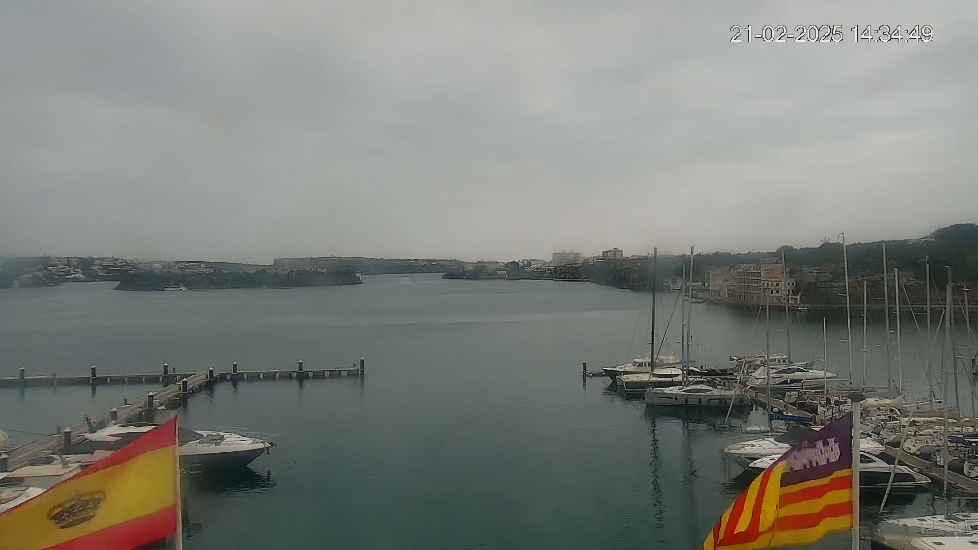 A marina filled with sailboats and motorboats is shown on an overcast day, with a town and hills visible across the water.