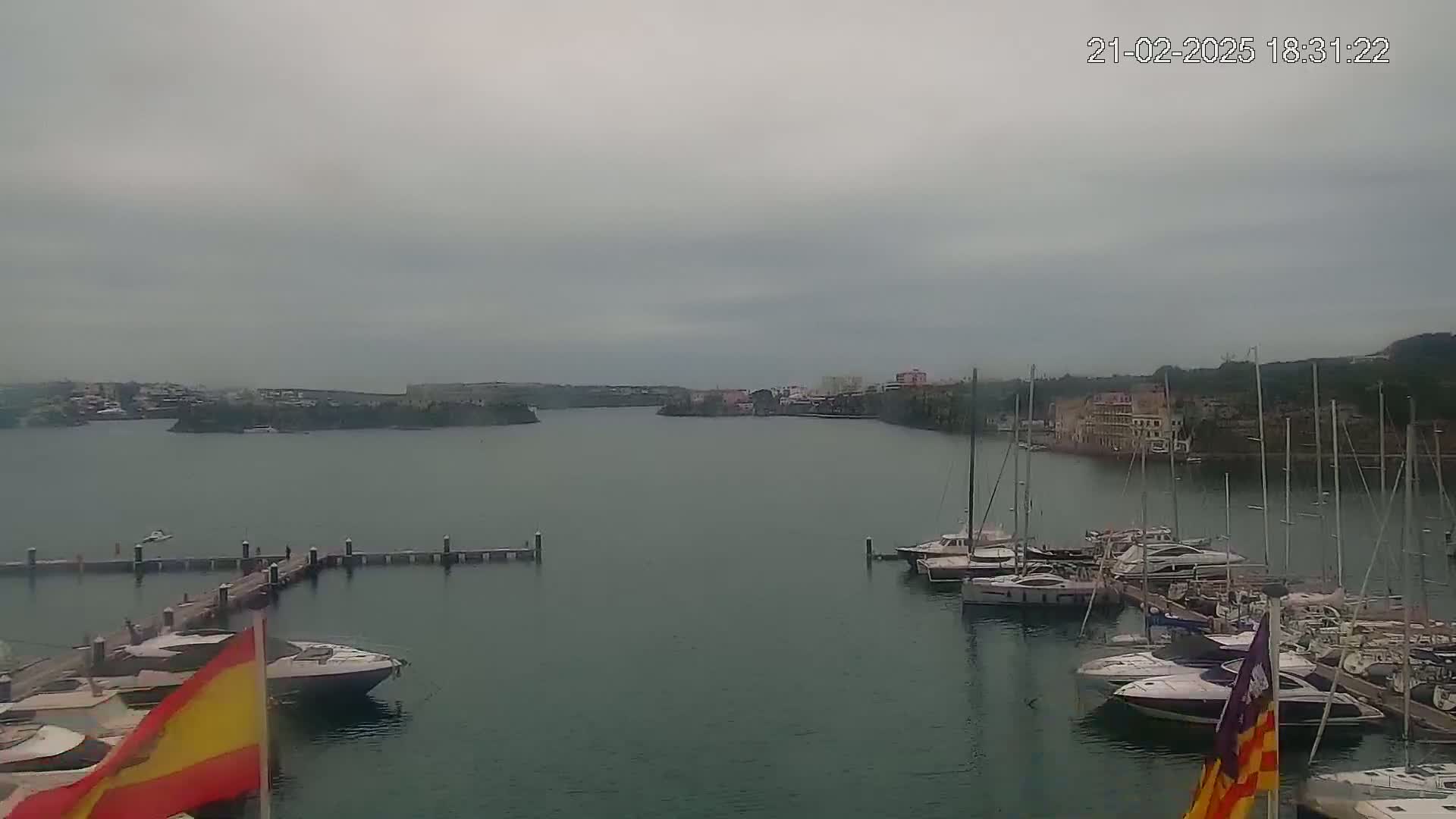 A marina filled with yachts and sailboats is seen on an overcast day, with a town and hills visible in the distance across calm water.