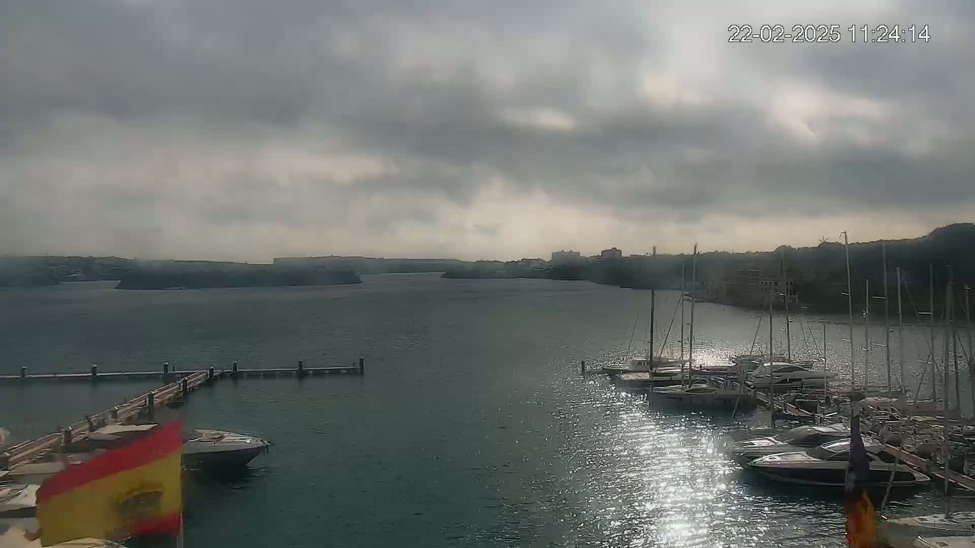 A marina filled with sailboats and yachts is shown on a partly cloudy day, with a calm body of water extending to a distant shoreline.