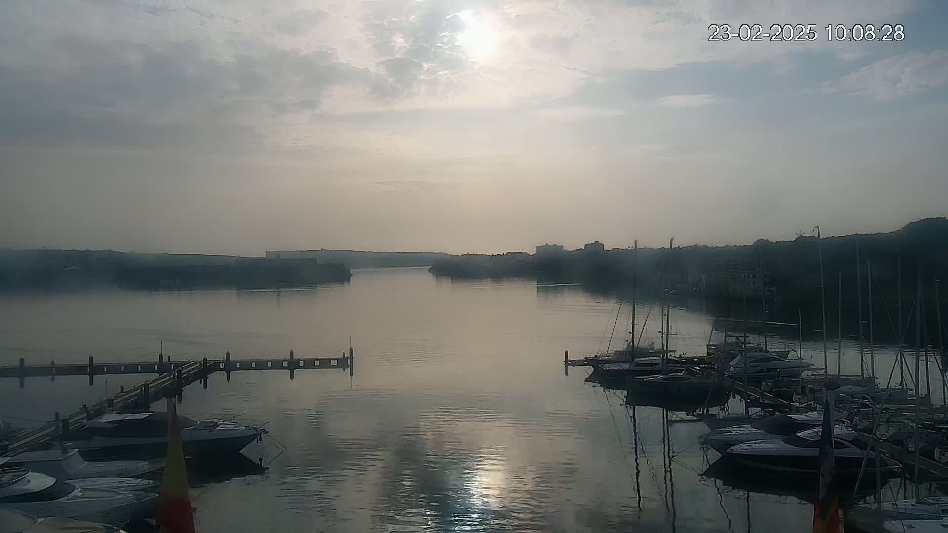A hazy sunrise illuminates a calm body of water where numerous boats are moored at a dock, with a distant shoreline visible.