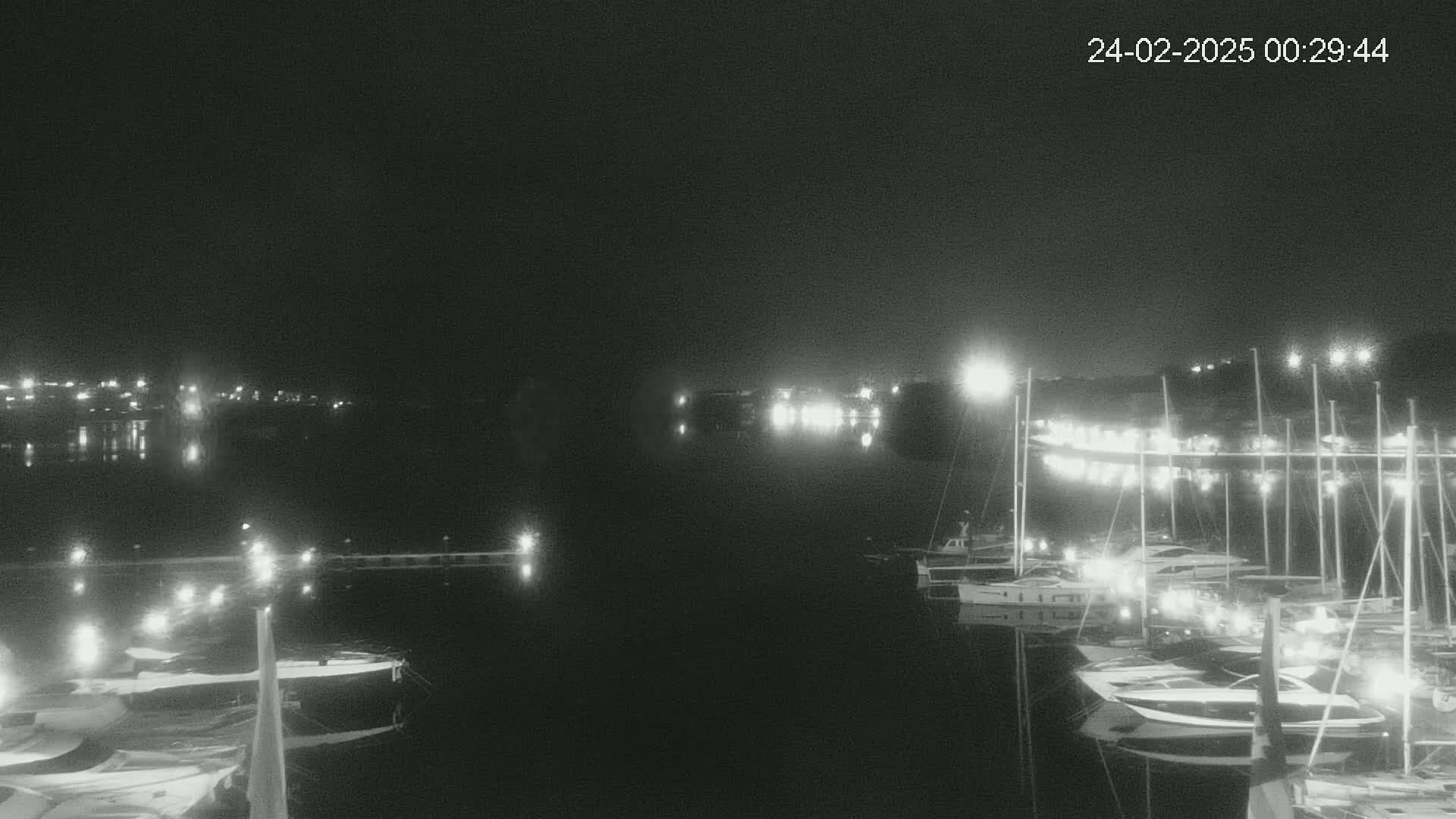 A nighttime, low-light view of a marina with numerous boats docked, illuminated by lights along the shoreline and on the boats themselves, reflecting on the calm water.