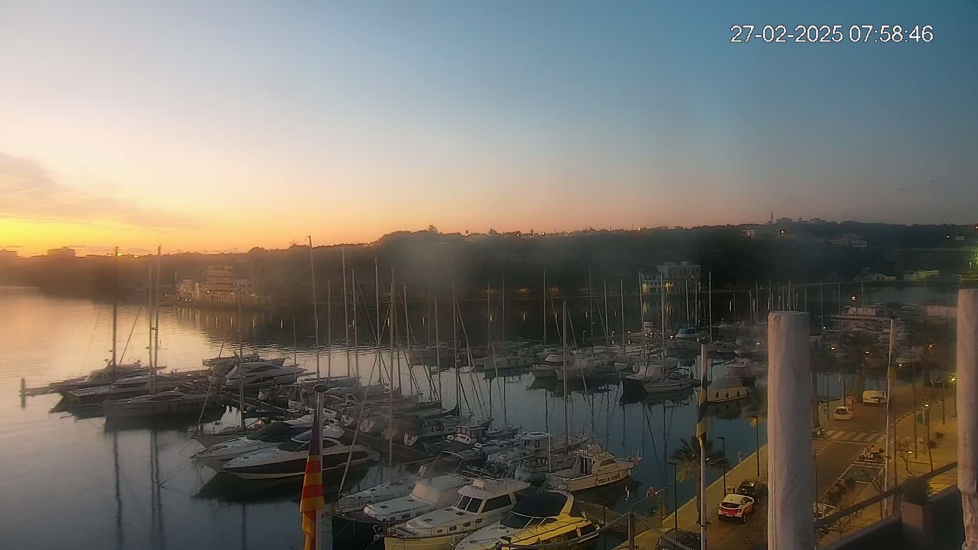 Many sailboats and motorboats are docked in a marina at sunrise, with calm water reflecting the colorful sky.