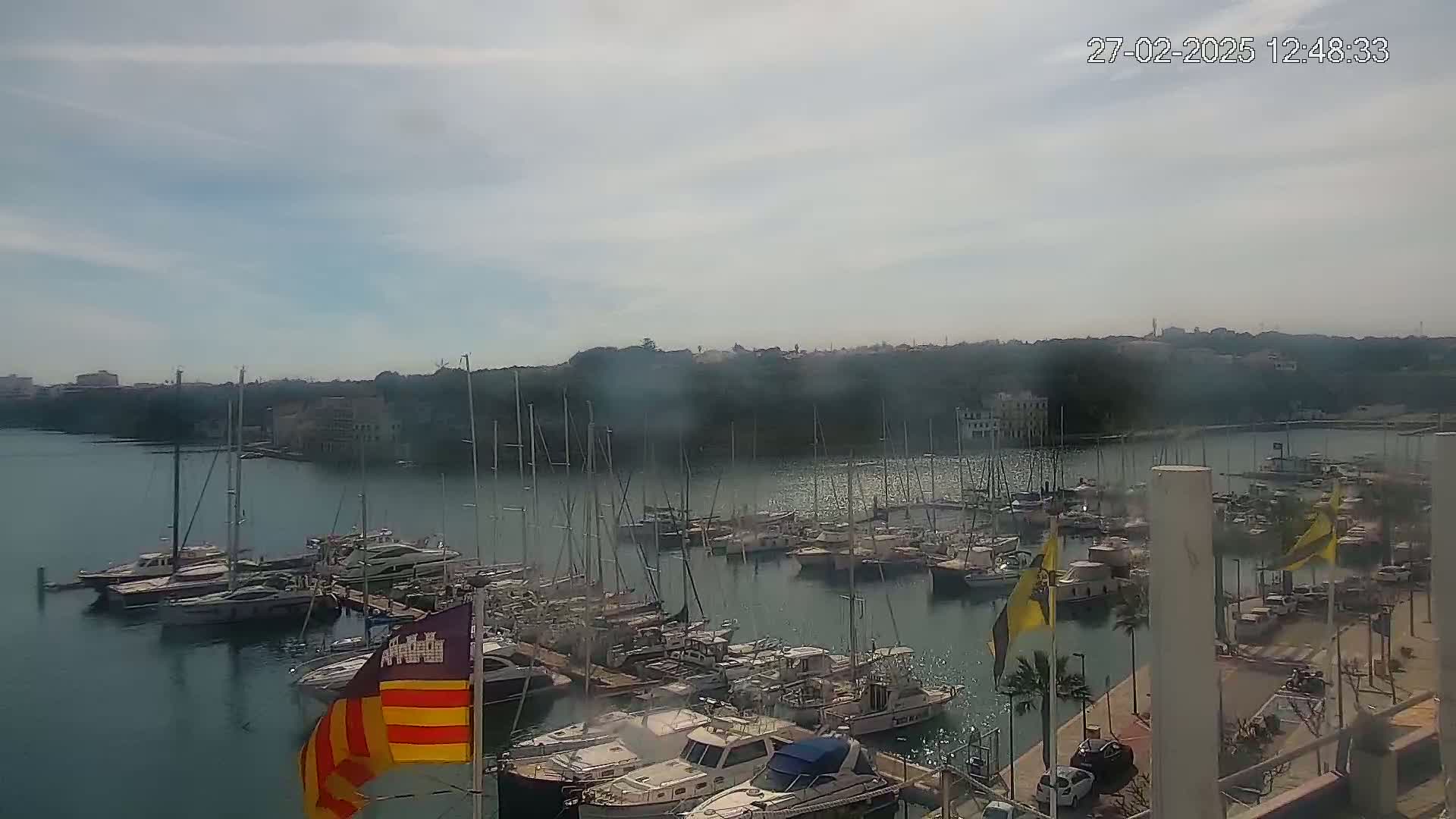 A marina filled with numerous sailboats and motorboats is situated on calm water under a partly cloudy sky.