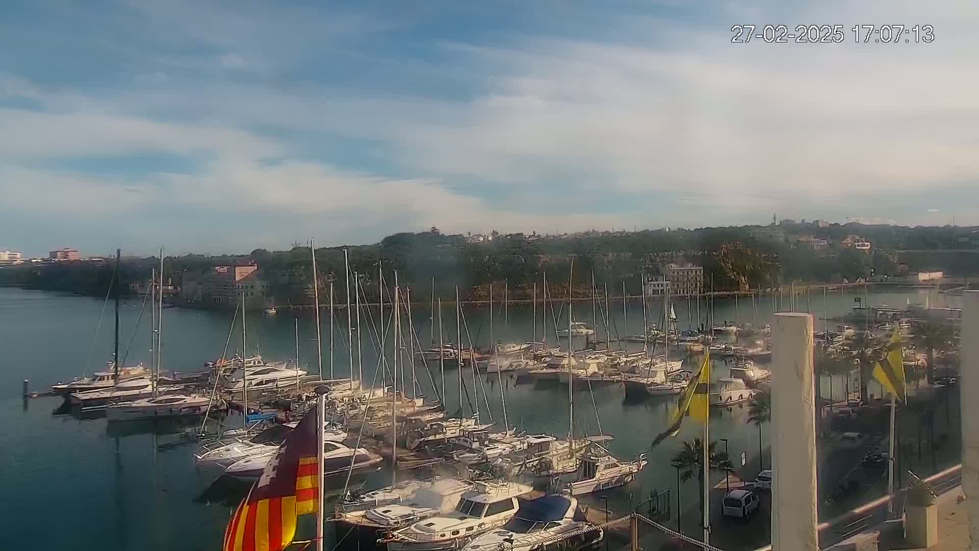 A marina filled with numerous sailboats and motorboats is situated on calm water under a partly cloudy sky.