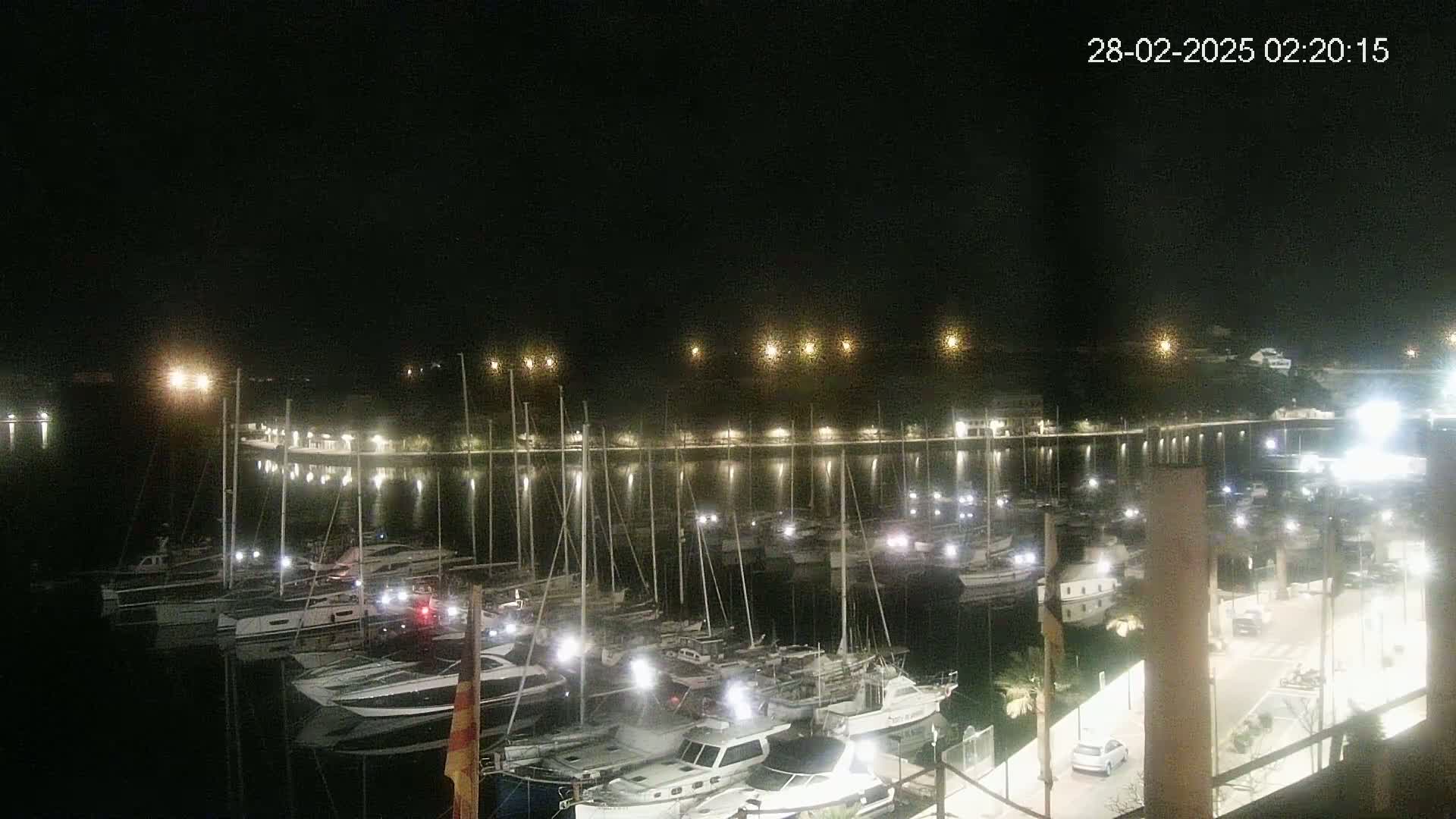 A nighttime view of a marina filled with sailboats and motorboats, illuminated by lights along a waterfront promenade.